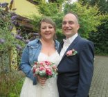 Brautpaar André und Marina Manuela Jung geb. Hecker Brautpaar vor Schmetterlingsflieder. Braut in weißem Kleid mit Jeansjacke und rosa Blumenstrauß. Bräutigam in blauem Anzug und weißen Hemd.